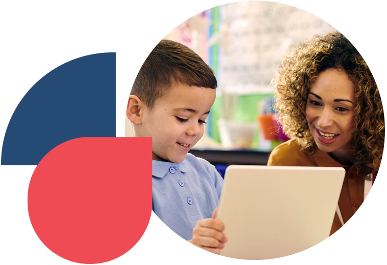 A smiling female teacher with curly hair leans in to look at a laptop screen alongside a young male student in a light blue polo shirt. The student is holding the laptop and smiling at the screen