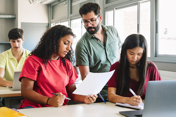 A male teacher with a beard and glasses stands over a desk, reviewing a document with two female students in a bright, modern classroom. One student, in a red shirt, holds the paper while the teacher points to the content; the other student, in a maroon shirt, takes notes. In the background, another student works on a laptop near a large window.
